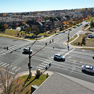 An aerial view of a suburban intersection with traffic lights and crosswalks. Several cars are stopped at the lights, and the surrounding area includes residential houses, landscaped yards, and trees with autumn foliage. The sky is clear, and the neighborhood appears well-maintained.